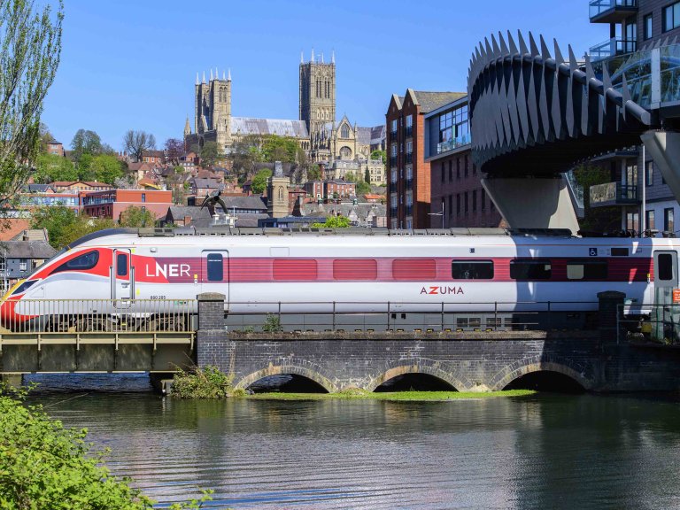 Chris Vaughan Photography - commercial photography | An LNER Azusa is pictured passing under a modern bridge in Lincoln city centre. In the foreground is water from the Brayford Pool reflecting the train, whilst Lincoln Cathedral is seen in the background.