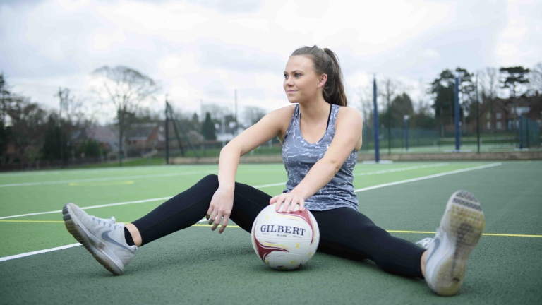 Chris Vaughan Photography - commercial photography | A netball player sits on a green coloured court, looking off camera. She is resting her hand on a netball in front of her.