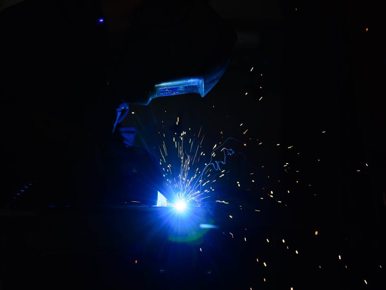 Chris Vaughan Photography - commercial photography | The blue flame from welding illuminates the mask worn by the worker. Sparks can be seen flying off in all directions against the black background.