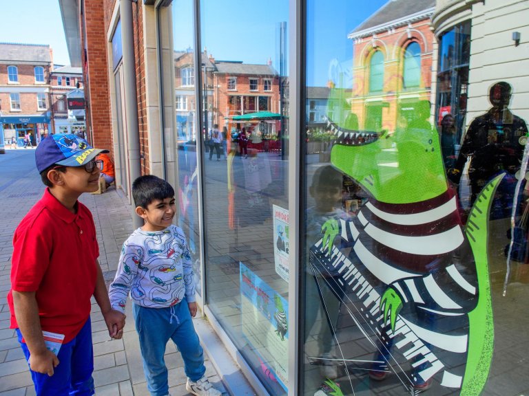 Two young children look at a window graphic of a green dinosaur, wearing a black and white stripy jumper, playing a keyboard.