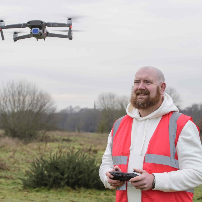 A profile picture of Chris Vaughan, owner of Chris Vaughan Photography, flying a drone. Chris is wearing a white hoodie and a high-vis jacket that has drone pilot written on the back.