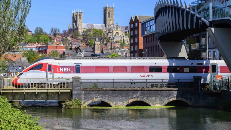 Chris Vaughan Photography - commercial photography | An LNER Azusa is pictured passing under a modern bridge in Lincoln city centre. In the foreground is water from the Brayford Pool reflecting the train, whilst Lincoln Cathedral is seen in the background.
