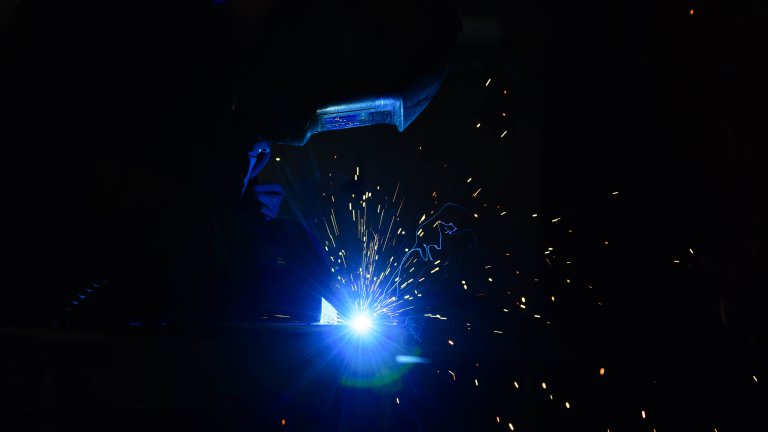 Chris Vaughan Photography - commercial photography | The blue flame from welding illuminates the mask worn by the worker. Sparks can be seen flying off in all directions against the black background.