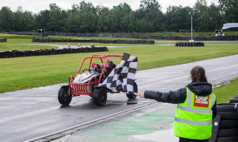 A GSF Car Parts colleague, wearing a branded yellow high-vis, waves a chequered flag as a motor kart crosses the finish line.