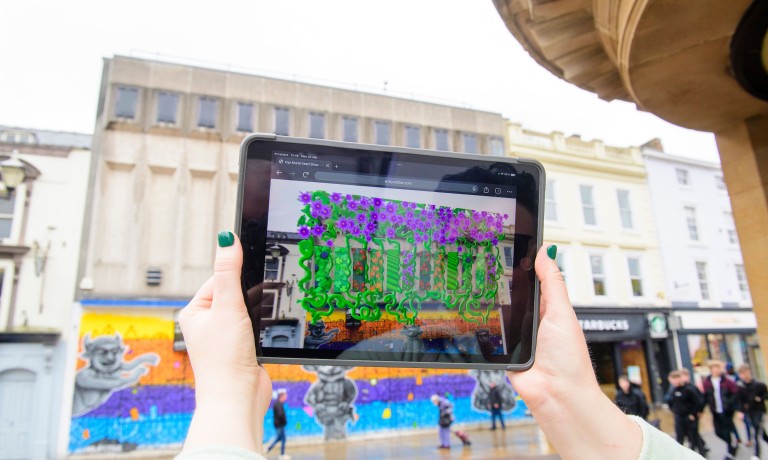 An artist holds an iPad which has digitally transformed the visual aspect of the building in the background.