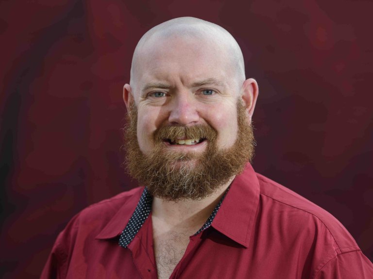 A profile picture of Chris Vaughan, owner of Chris Vaughan Photography. Chris is wearing a red shirt, and is photographed near a red brick wall.